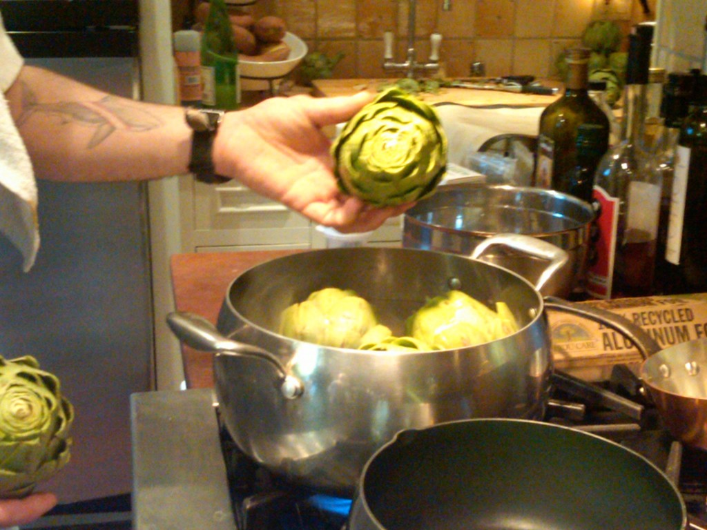 PLACE THE ARTICHOKES IN SALTED ACIDULATED SIMMERING WATER. Acidulated water is water where some sort of acid is added—often lemon juice, lime juice, or vinegar—to prevent cut or skinned fruits or vegetables from browning so as to maintain their appearance.