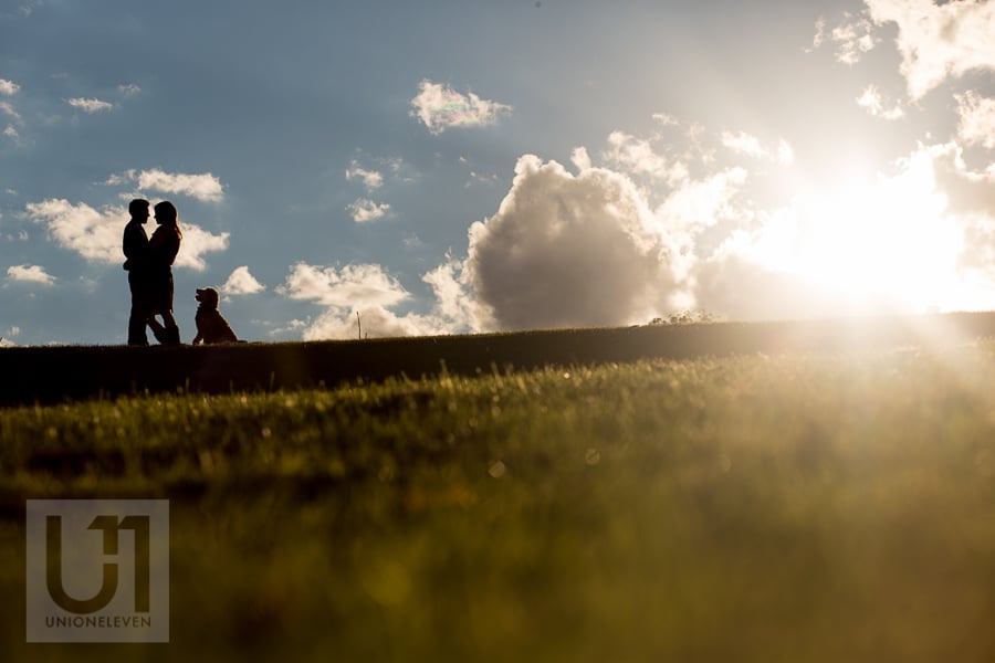 Silhouette of young couple standing in an embrace on a hill, with their dog sitting and facing them on the right.