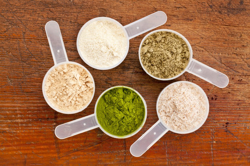 five plastic measuring cups of different superfood supplement powders (form bottom clockwise: wheatgrass, maca root, whey protein, hemp seed protein, psyllium husk) on grunge wood background