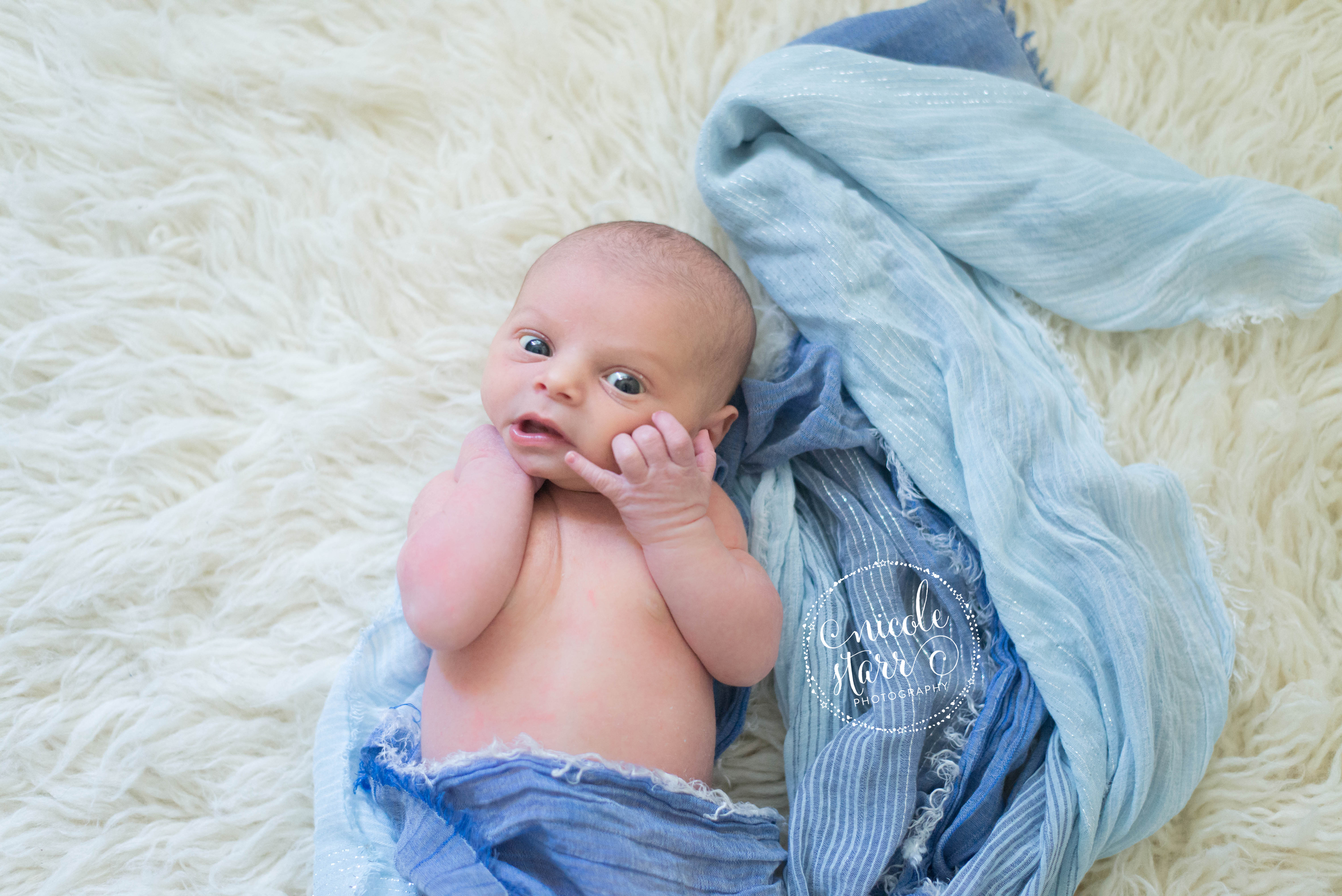 wide eyed newborn baby boy on a flokati rug with a blue wrap