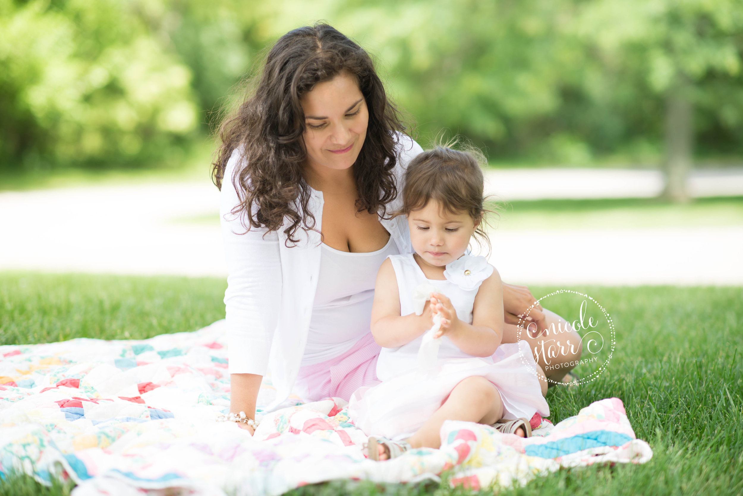 mommy and baby girl in a park on a quilt reading a book