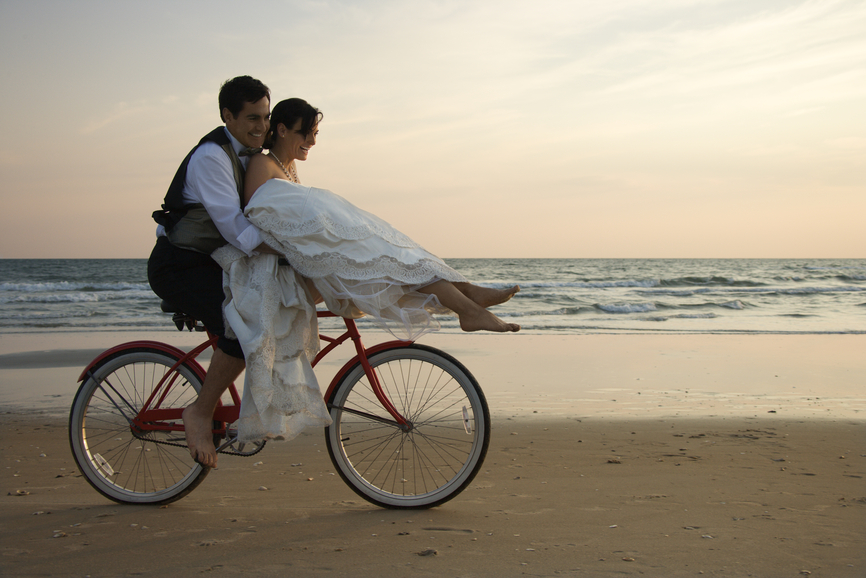 Bride rides the handle bars of a bicycle being driven by her groom on beach. Horizontal shot.