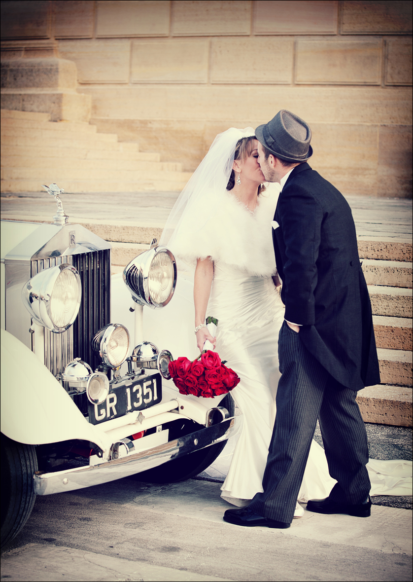 A Bride and Groom kiss next to a vintage car in front of the Philadelphia Museum of Art.