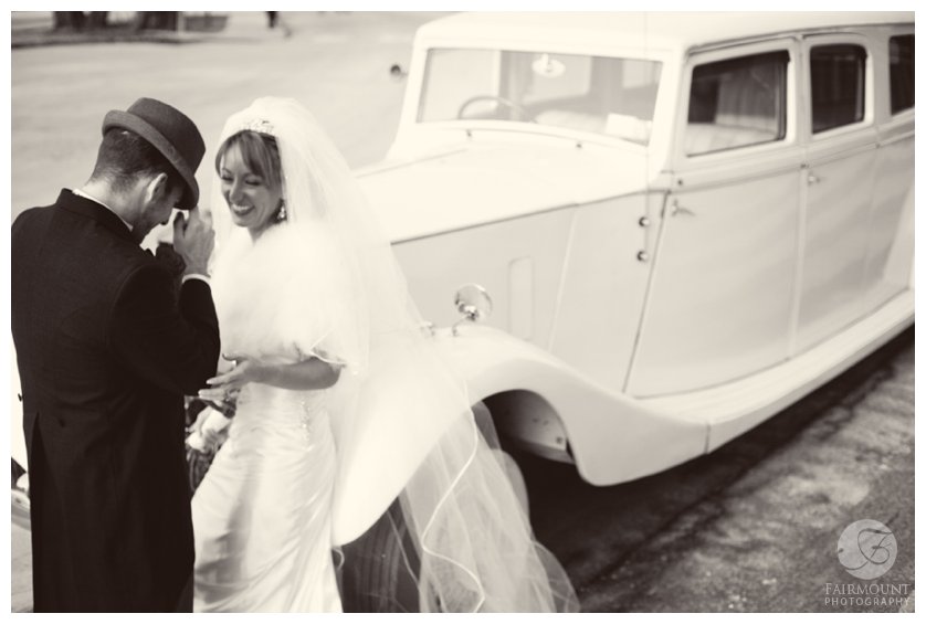 groom in fedora tips hat to bride leaning on antique car