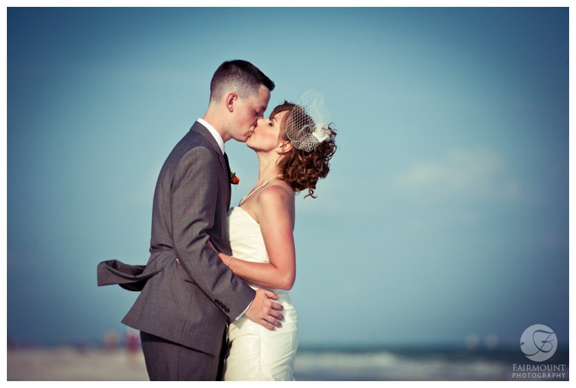 vintage-styled portrait of bride in birdcage veil and strapless dress kissing groom in gray suit on the beach