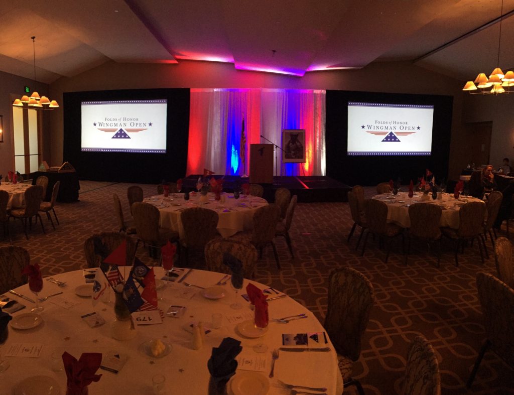 Picture of AV for You Folds of Honor Wingman Open equipment set-up featuring two projectors, two 13' truss towers, with six etc fours lights, and 16' drape behind the stage with 8 up lights set to red, white, and blue.