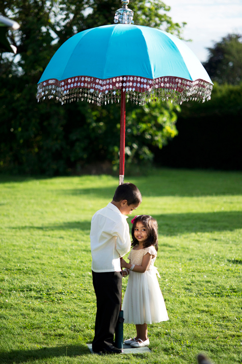 Exterior shot of young boy and girl standing under a turquoise parasol on the lawn