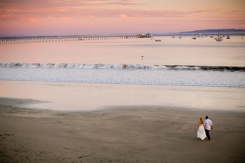 Avila Beach Engagement pictures of bride and groom walking on beach during pink sunset.