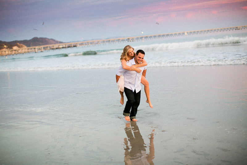 Avila Beach Engagement pictures of bride on groom's back running on beach during pink sunset.