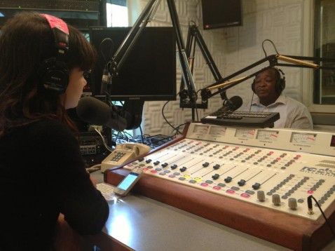 the back of a young woman interviewing an African man across a sound board inside a radio station