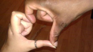 Two hands forming a heart with their fingers, one light brown with a ring on the thumb and long nails, the other darker brown with short nails.  These are Patty and Leroy's hands.