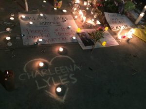 Image taken by Lisa Ganser at a vigil for Charleena Lyles, in Seattle, WA, 6/18/17. Image description: a sidewalk at night with multiple votive candles and tall candles illuminating warm light in the darkness. Signs and flowers are spread throughout the sidewalk. A chalk drawing of a heart with 'Charleena Lyles' in the center. One white poster says: "Black Lives Matter/People with Mental Illness Matter" 