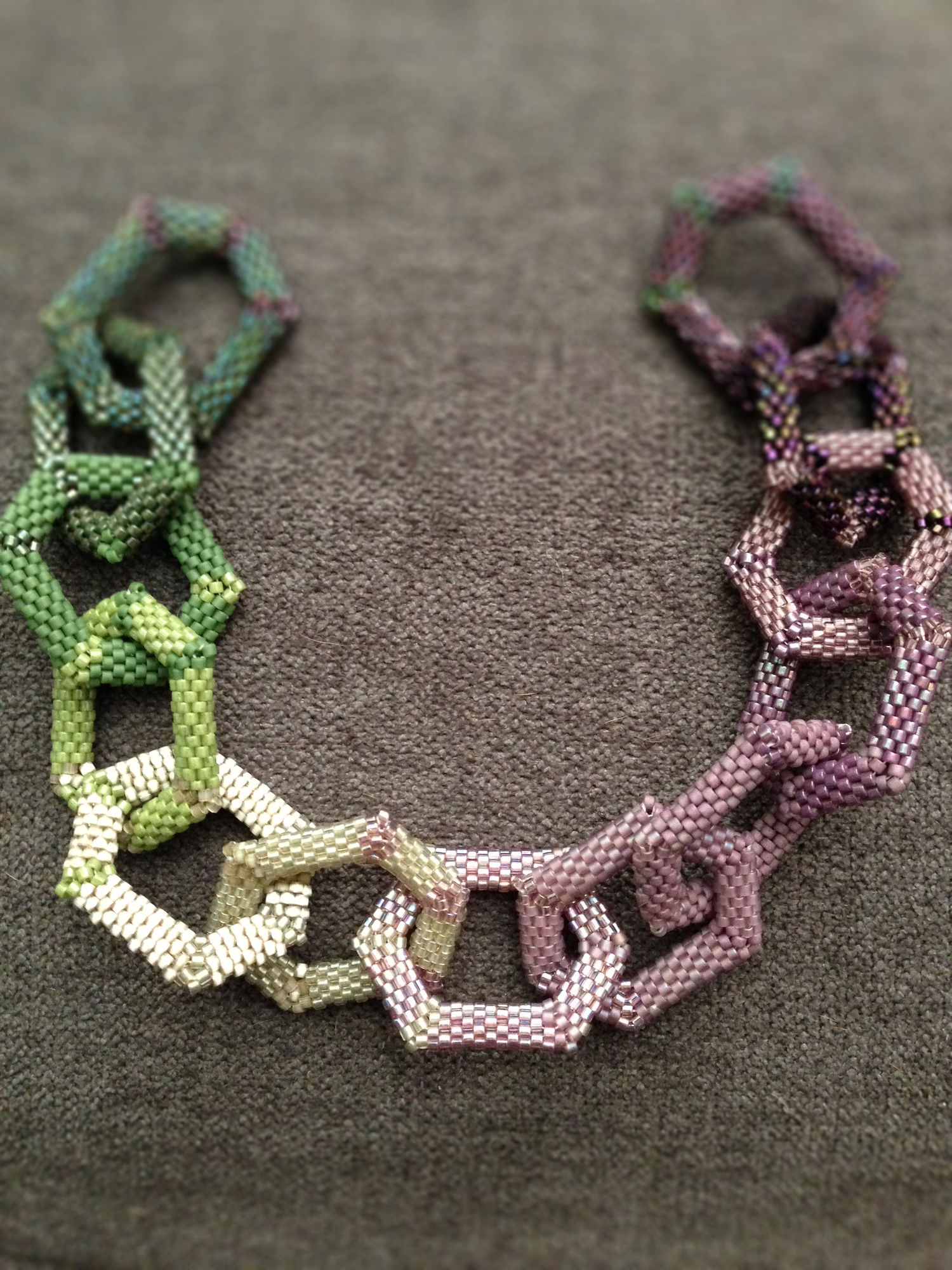 A string of peyote hexagons in a spectrum from dark green to light green to pale silver to lavender to dark purple.