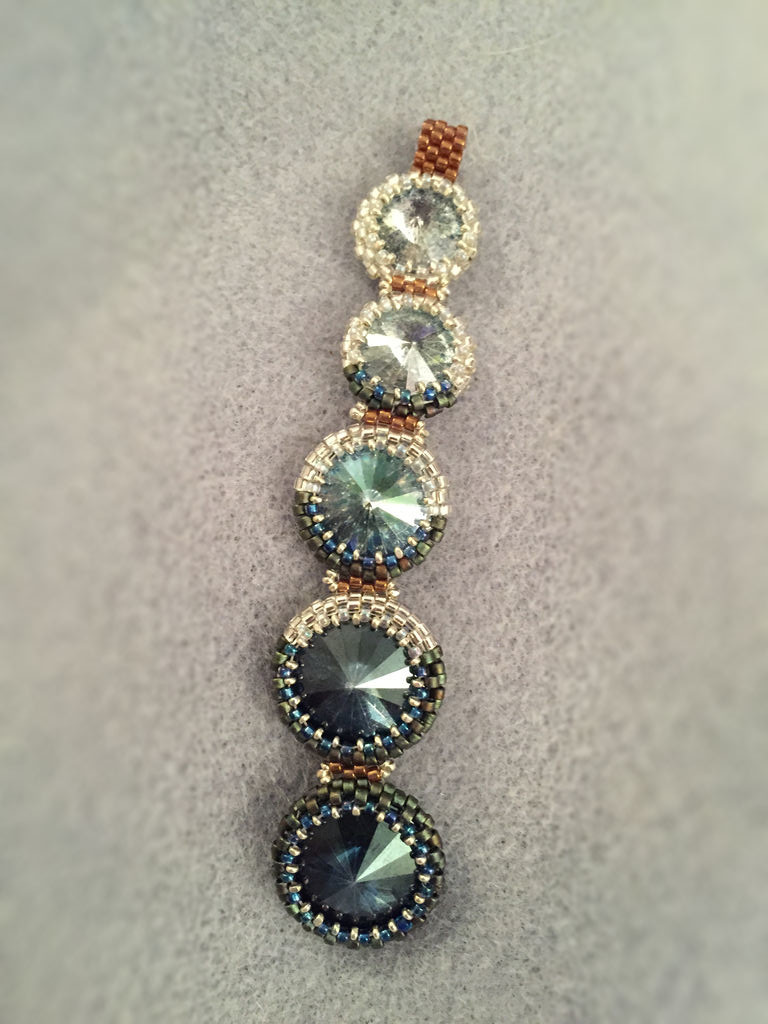Pendant made from a series of Swarovski rivolis in dark and pale blue. Silver and blue beaded bezels mimic the progression of the phases of the moon, and bronze peyote strips connect the rivolis to one another in series.