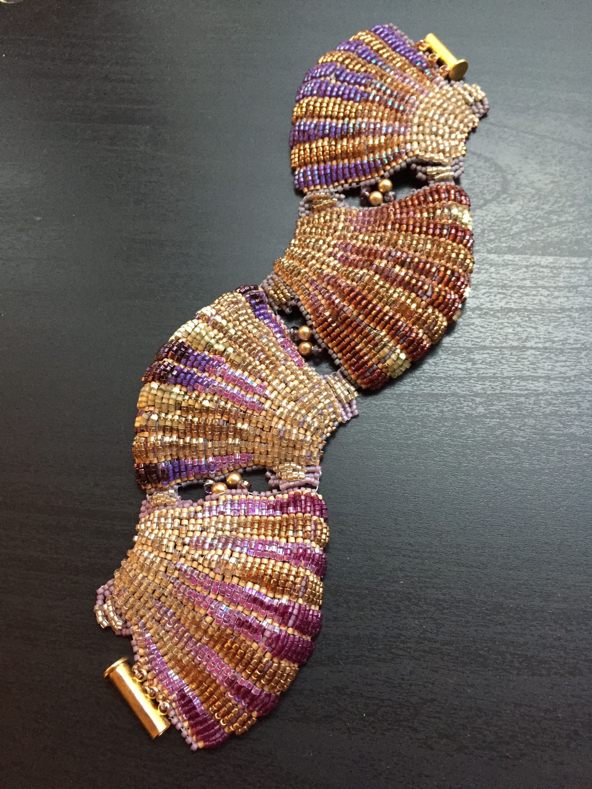 A cuff bracelet made of four beaded shell shapes, placed in the shape of a wave. Strong purples and bright golds throughout.