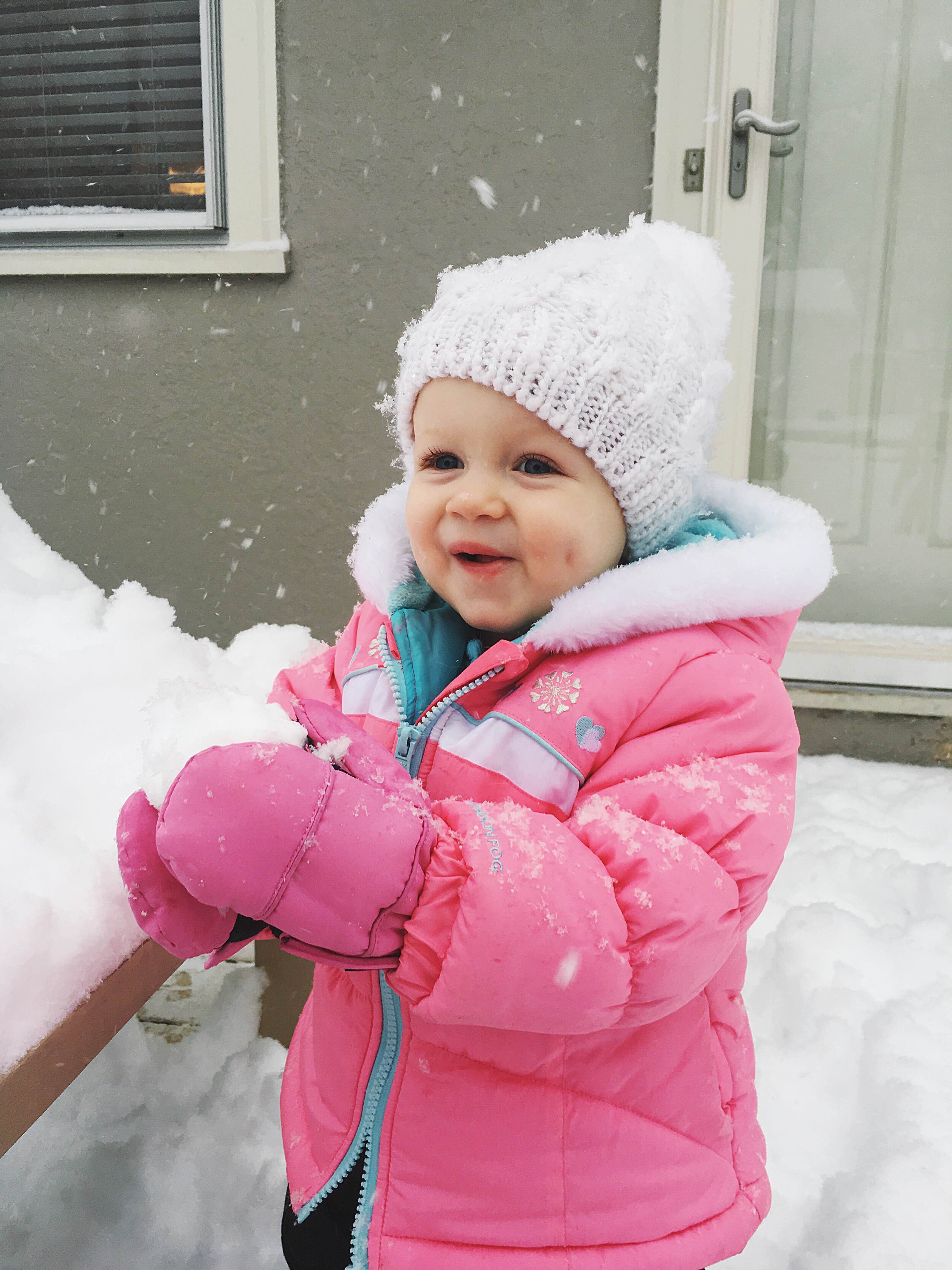 toddler playing in the snow in pink snowsuit and white pom pom hat