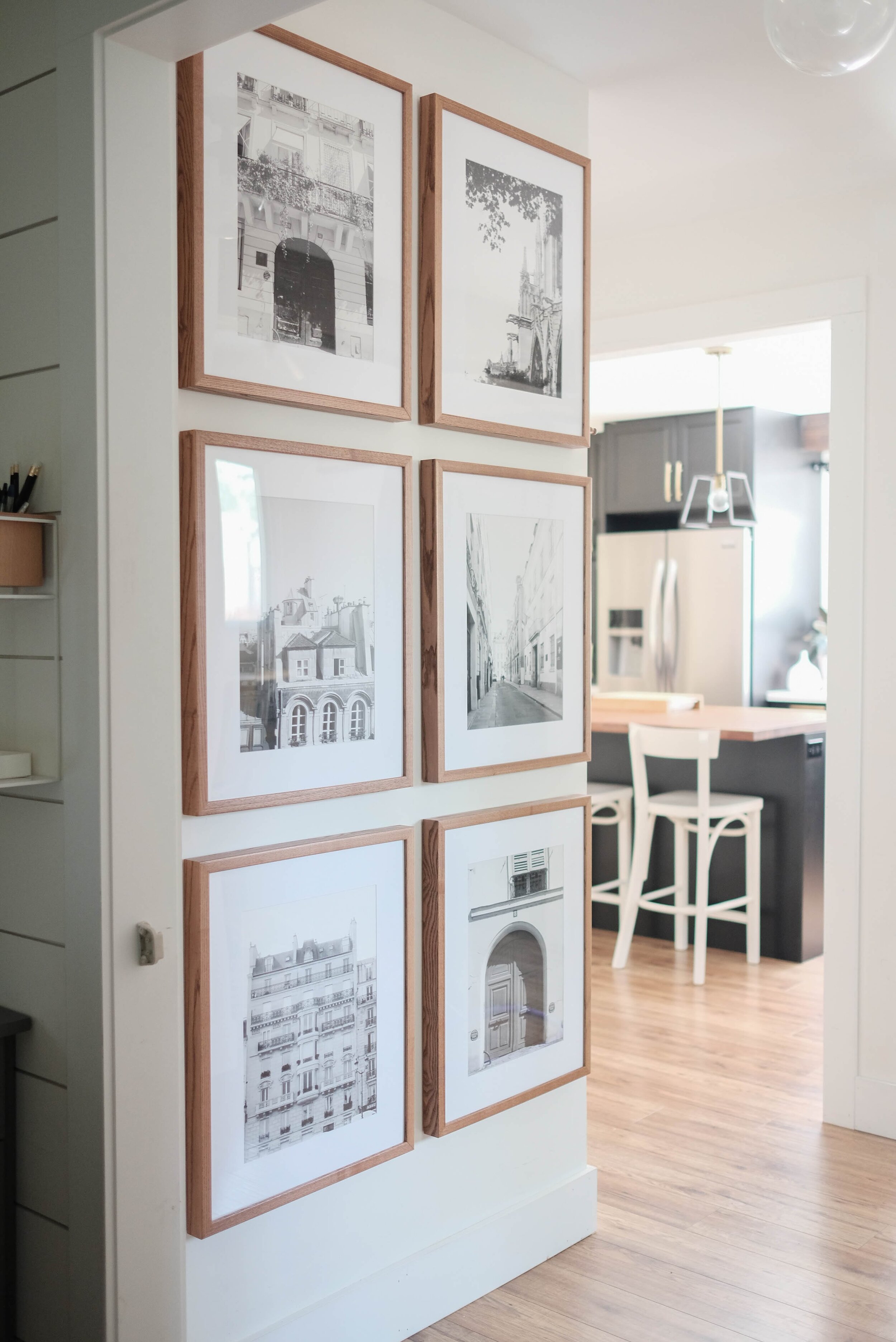 Symmetrical gallery wall in a hallway with light walnut frames from PosterJack. View of a kitchen with black cabinets to the right.