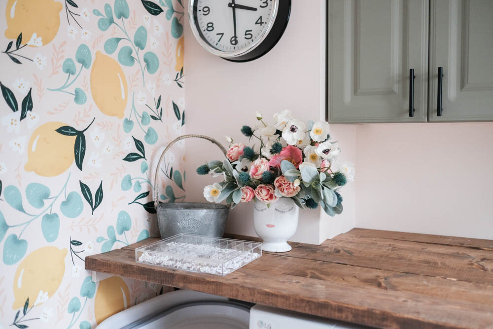 close-up of laundry room countertop with lemon wallpaper, fake flowers, green cabinets and pink walls