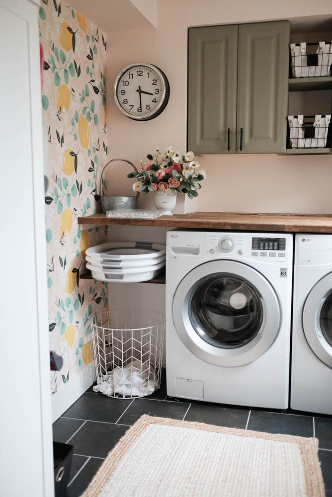 laundry room with pink walls and lemon wallpaper, stacked laundry baskets, green cabinets, and grey slate tiles on the floor
