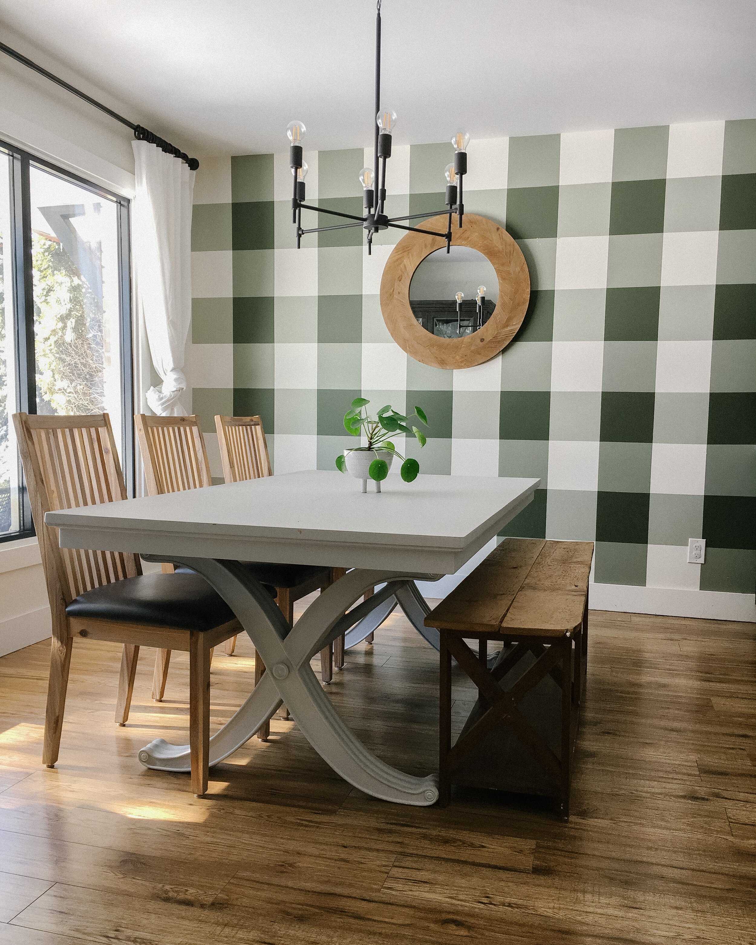 dining room with green buffalo plaid wall, grey table, 3 wooden chairs and a wooden bench, round wooden mirror on the wall and black chandelier.