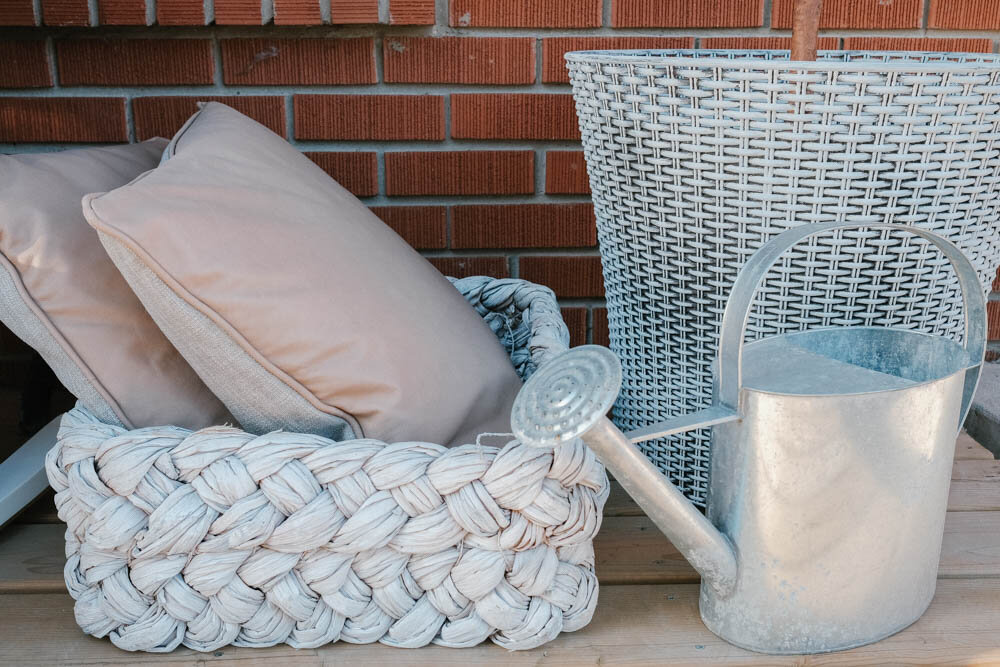 Tress white washed tray and Belez yuma mink taupe cushions from Article sitting on a front porch. Pillows are inside the basket and next to them is an aluminum watering can and a white washed flower pot.