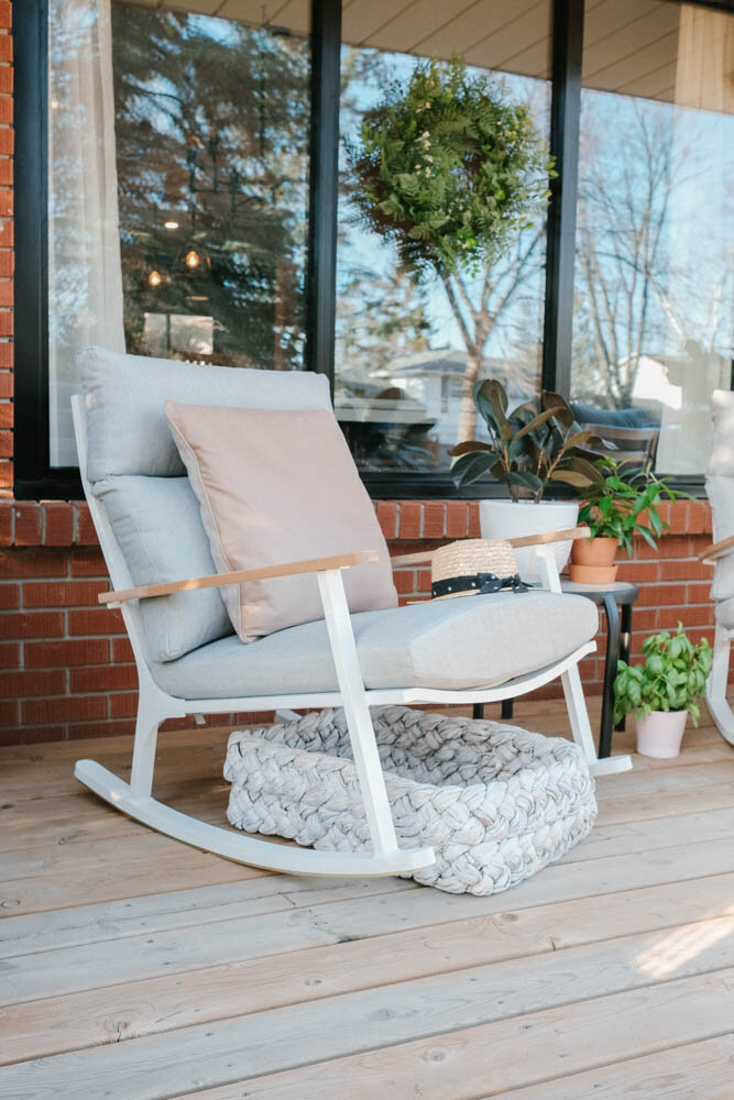 Modern white and grey rocking chair with a taupe pillow on it and white basket underneath. Porch has wood floors and red brick walls, there's a wreath hanging on the window and plants on a small table and on the floor.