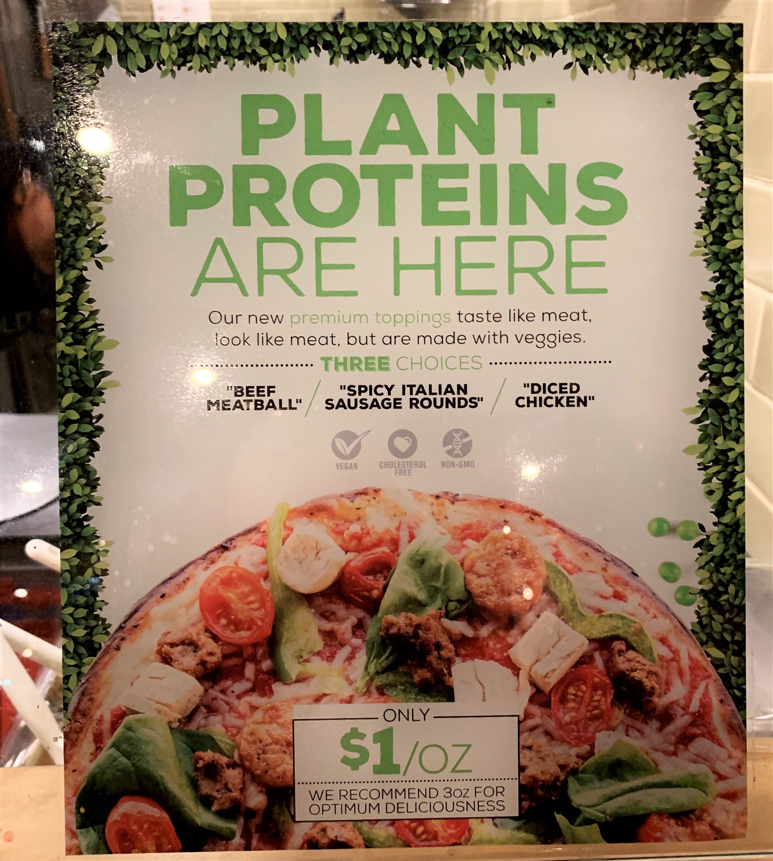A sign at Pieology Pizzeria that says "plant proteins are here" including beef meatballs, spicy Italian sausage, and diced chicken. It's also tagged as vegan, cholesterol free, and non-gmo.