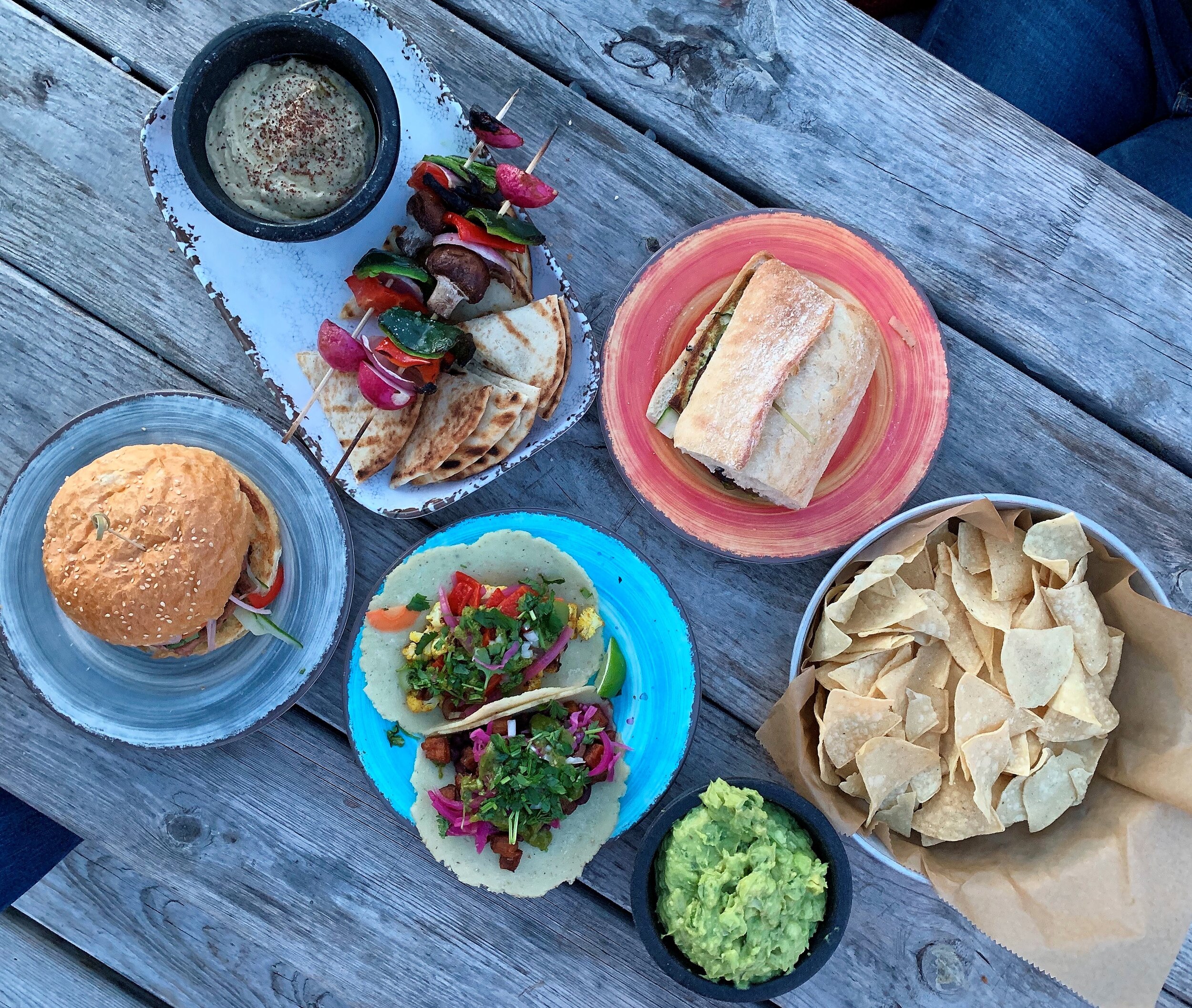 A picnic table full of vegan food including banh mi, tacos, guacamole, and vegetable skewers