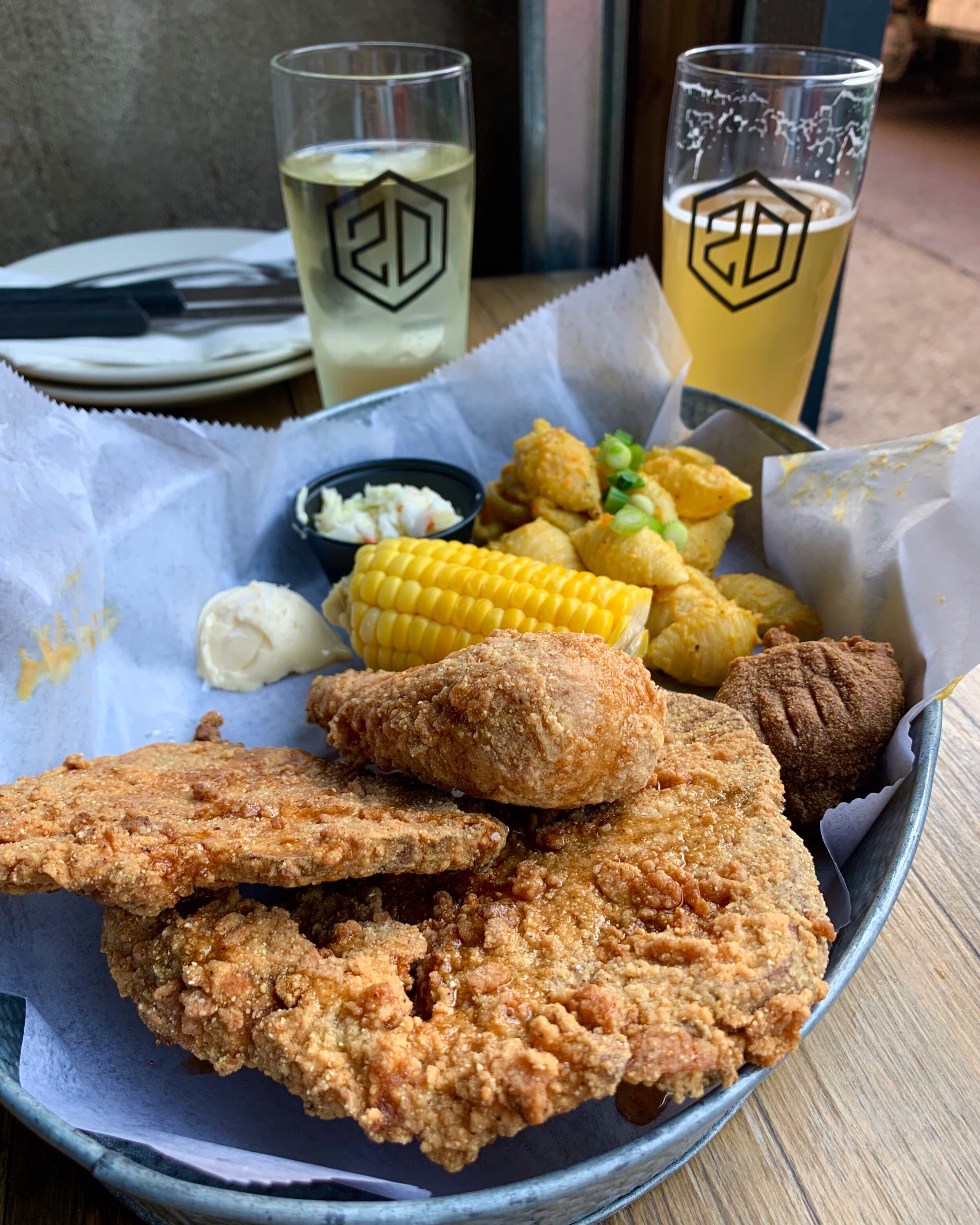 A basket of fried vegan chicken, corn on the cob, and mac and cheese with two glasses of beer in the background