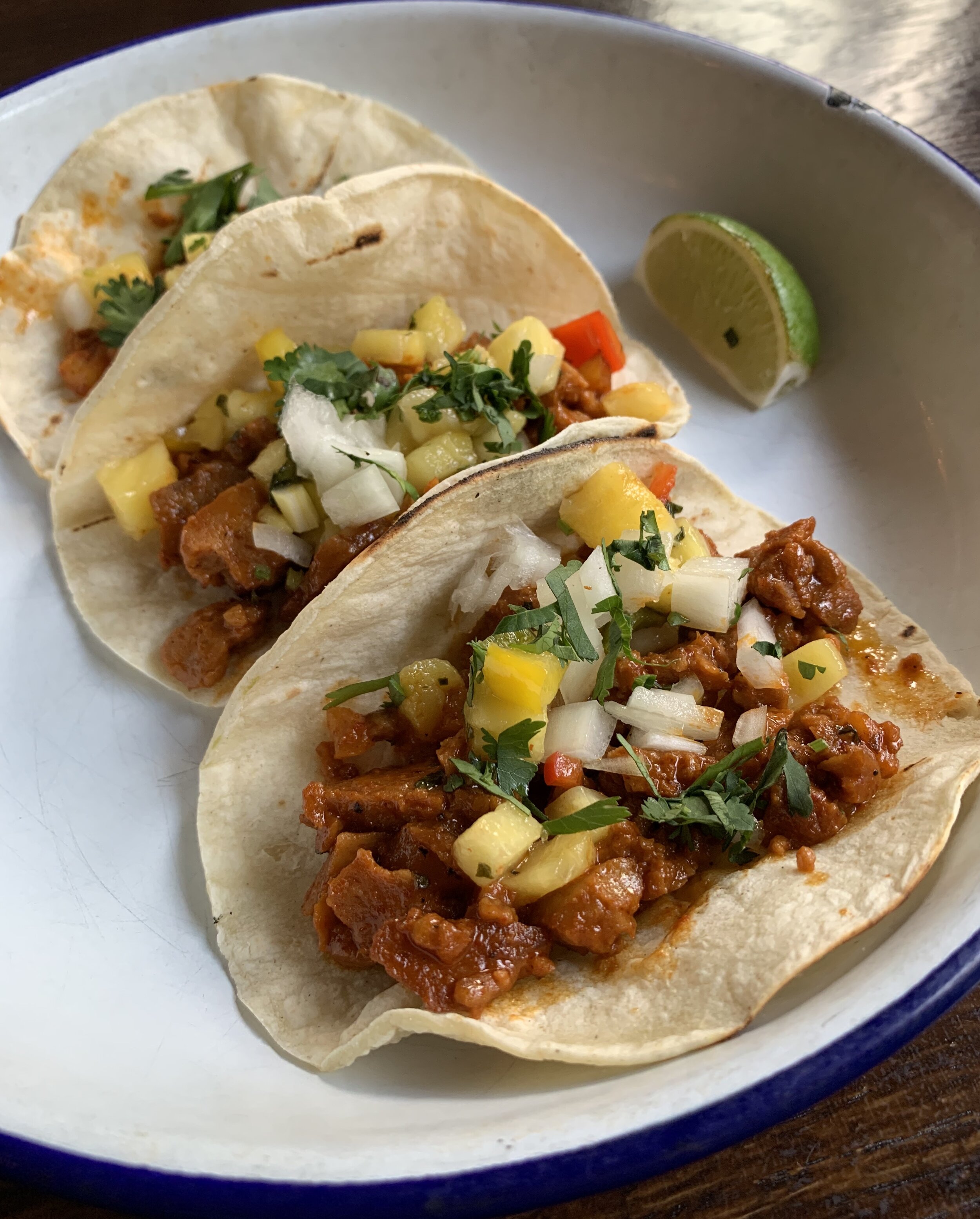 three tacos with sauced seitan, onions, and pineapple salsa with a lime wedge off to the side