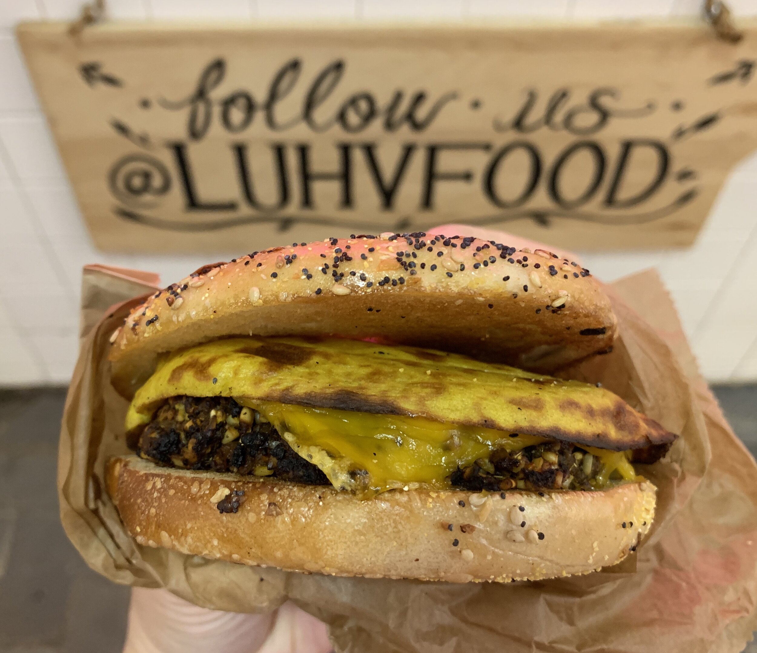 An everything bagel with vegan sausage and cheese, plus tofu with a sign in the background that reads "follow us @luhvfood"