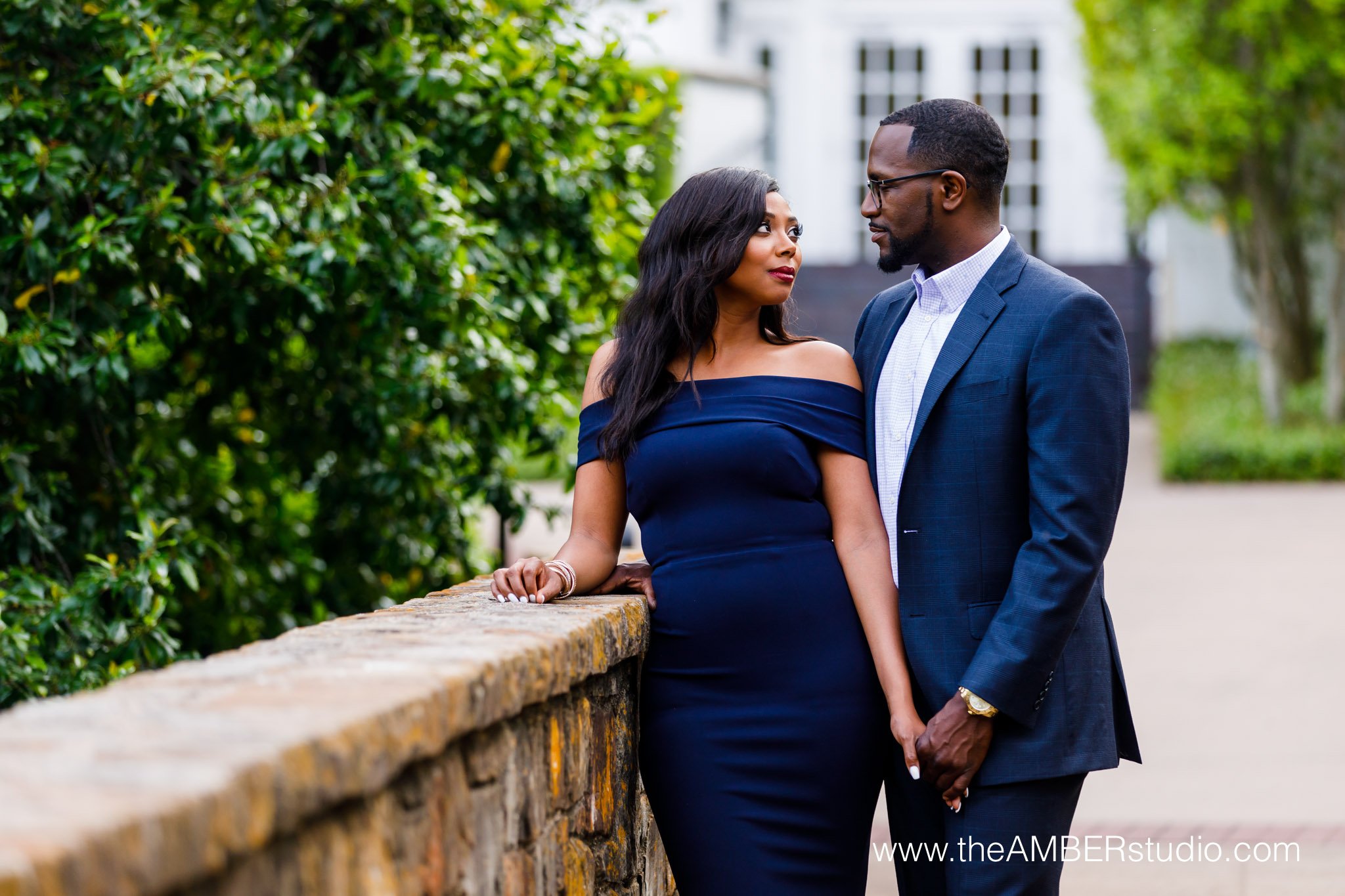 Black couple at their engagement session in Dallas at Arlington Hall. She is wearing a floor length navy gown, and he is wearing a blue suit with a white shirt unbuttoned at the top. They are looking at each other. 
