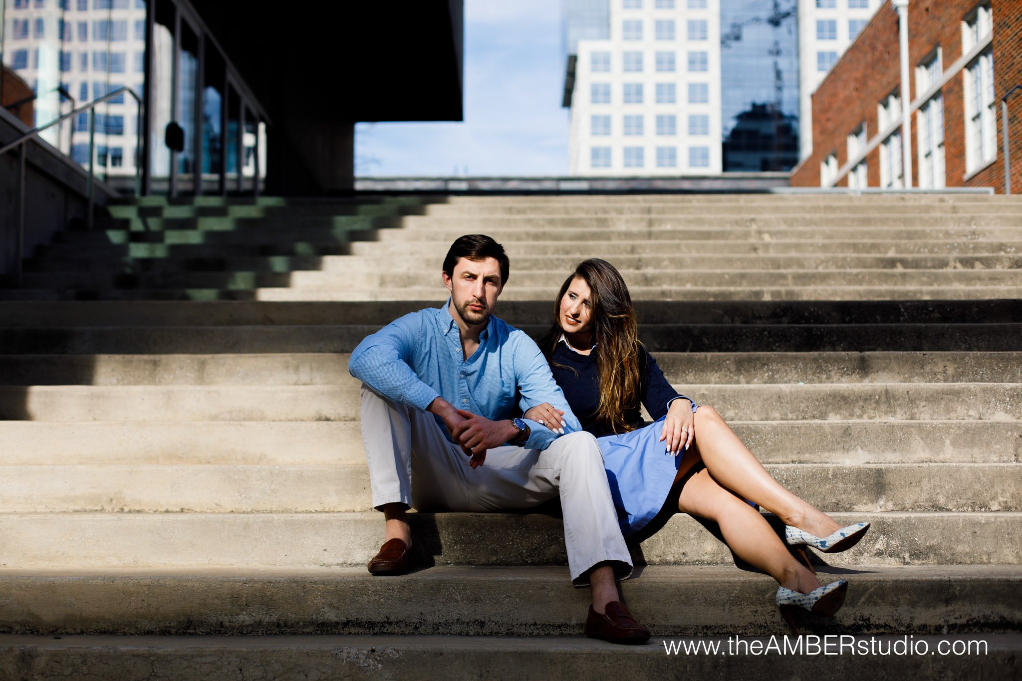 Multicultural couple at their Dallas engagement session in the Arts District. They are both sitting on the stairs in the sunlight. He is looking at the camera, and she is looking at him.