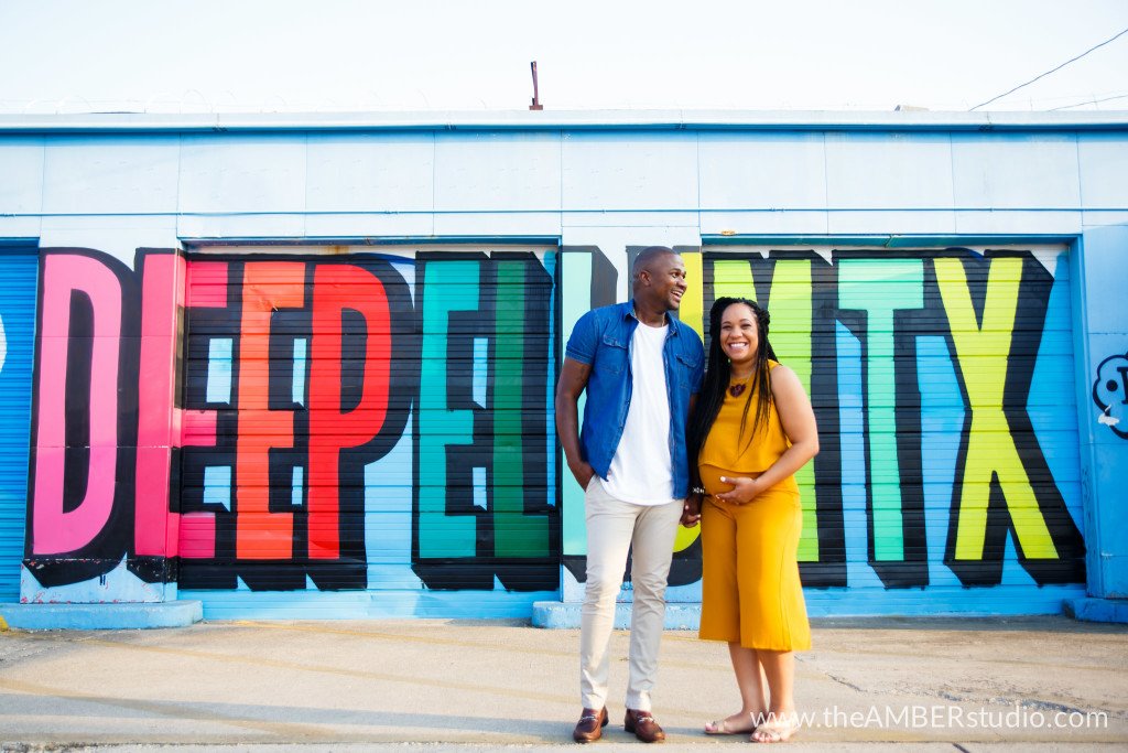 Black couple at their maternity session in the Deep Ellum neighborhood of Dallas. They are laughing and holding hands and standing in front of a colorful mural. She is wearing a yellow jumpsuit.