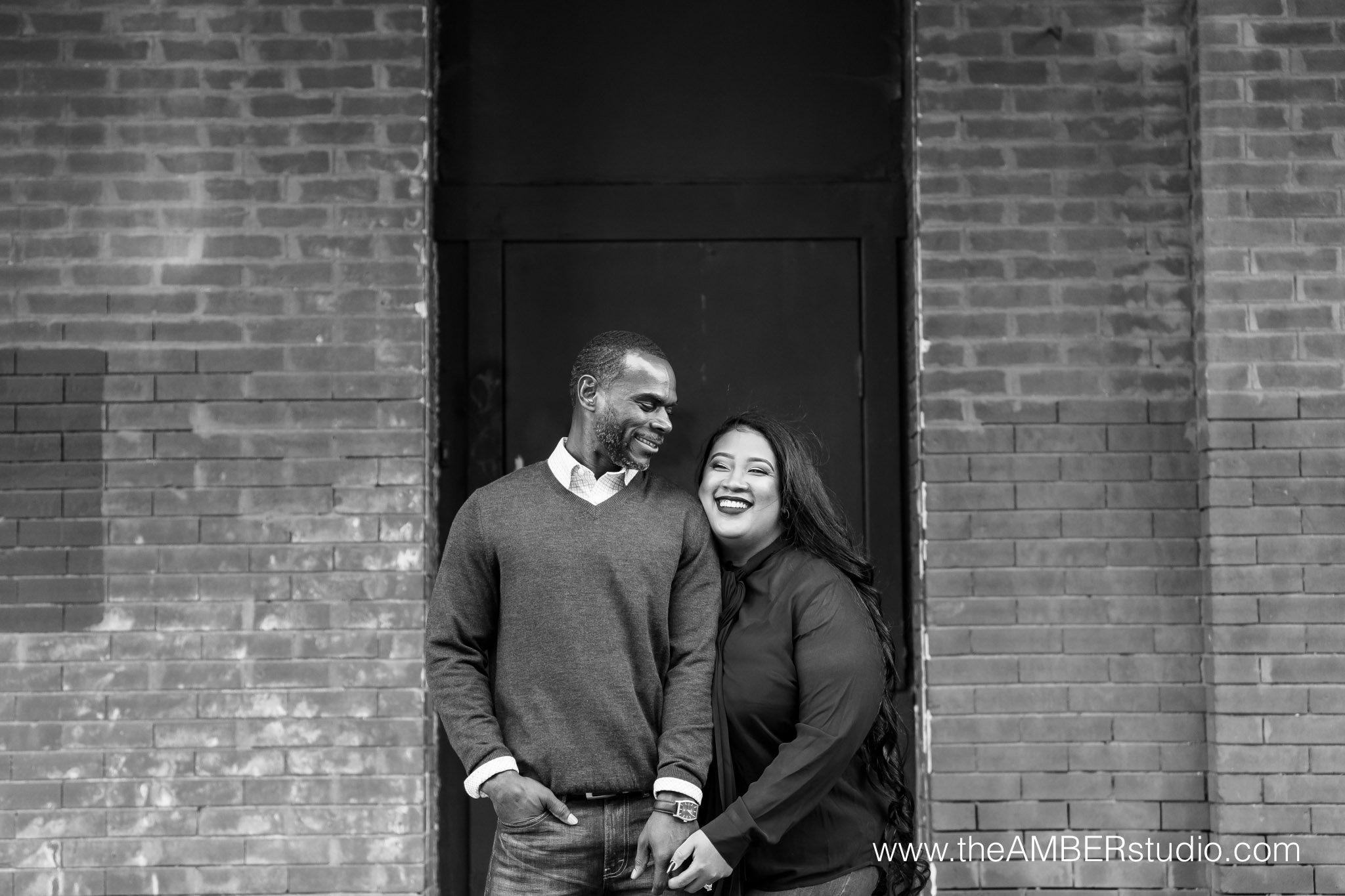 Black & white image of multicultural couple at the engagement session in downtown Dallas. They are laughing and holding hands in front of a brick wall.