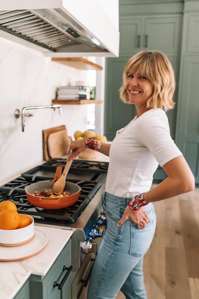 Our Place by popular Indianapolis lifestyle blog, Seersucker and Saddles: image of a woman wearing a white t-shirt and jeans and cooking at her gas stove with an orange Our Place Always Pan. 