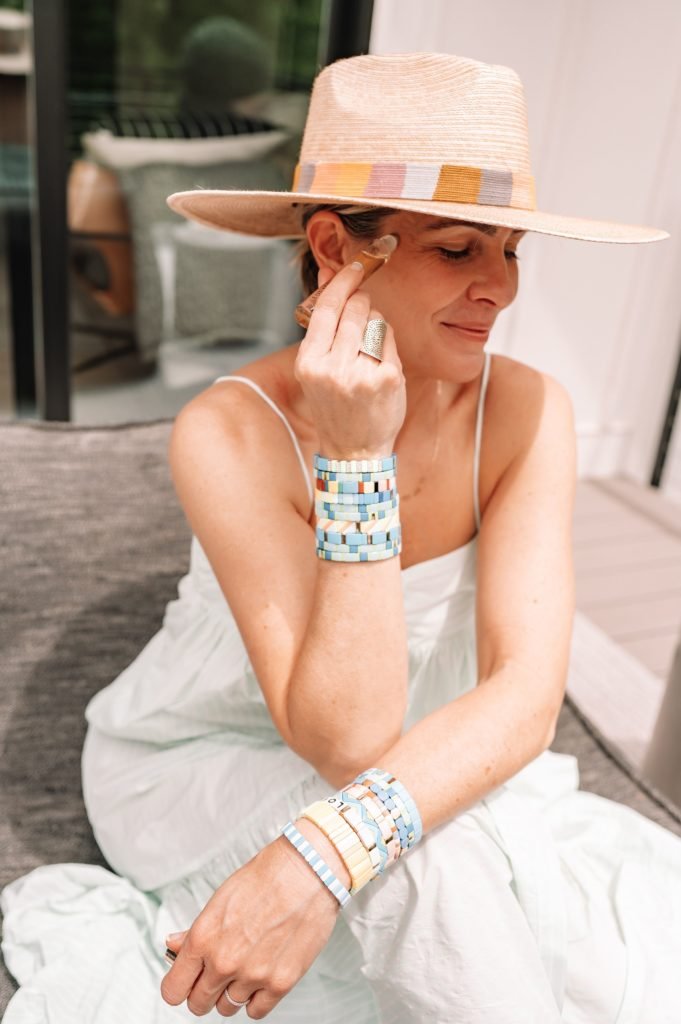 Equilibria Memorial Day Sale by popular Indianapolis lifestyle blog, Seersucker and Saddles: image of a woman wearing a straw fedora and light blue ruffle hem maxi dress while apply Equilibria CBD to the temple of her head. 