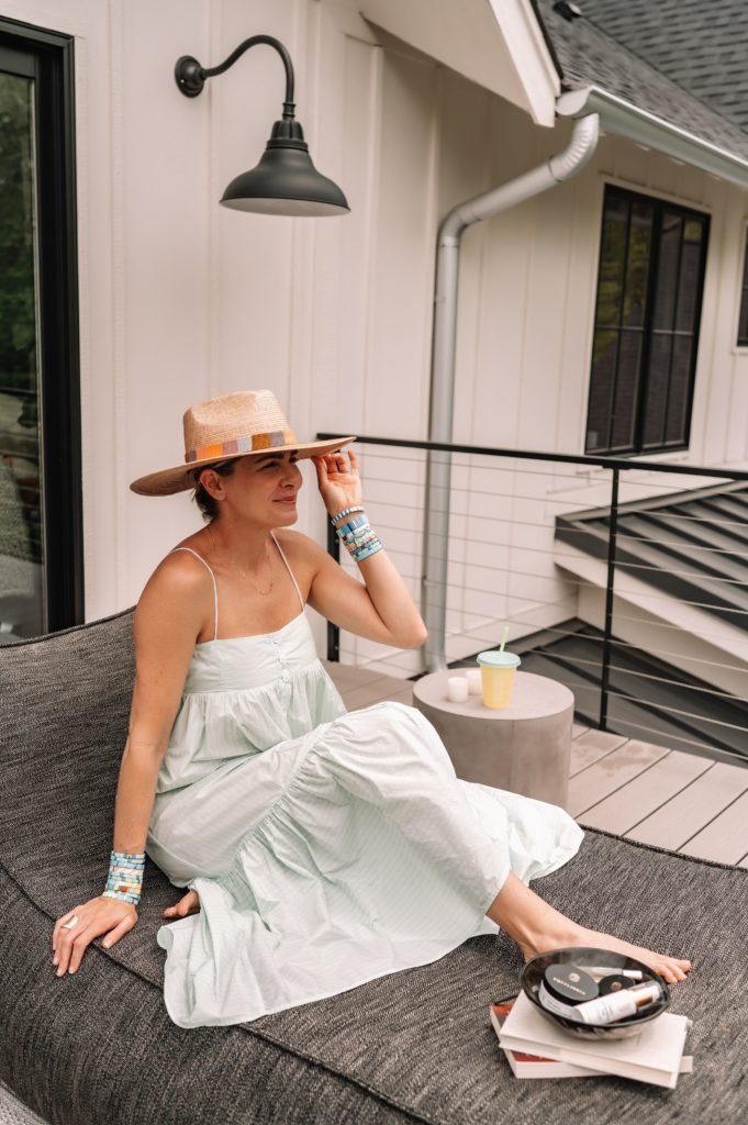 Equilibria Memorial Day Sale by popular Indianapolis lifestyle blog, Seersucker and Saddles: image of a woman wearing a straw fedora and light blue ruffle hem maxi dress while sitting outside next to some Equilibria products. 