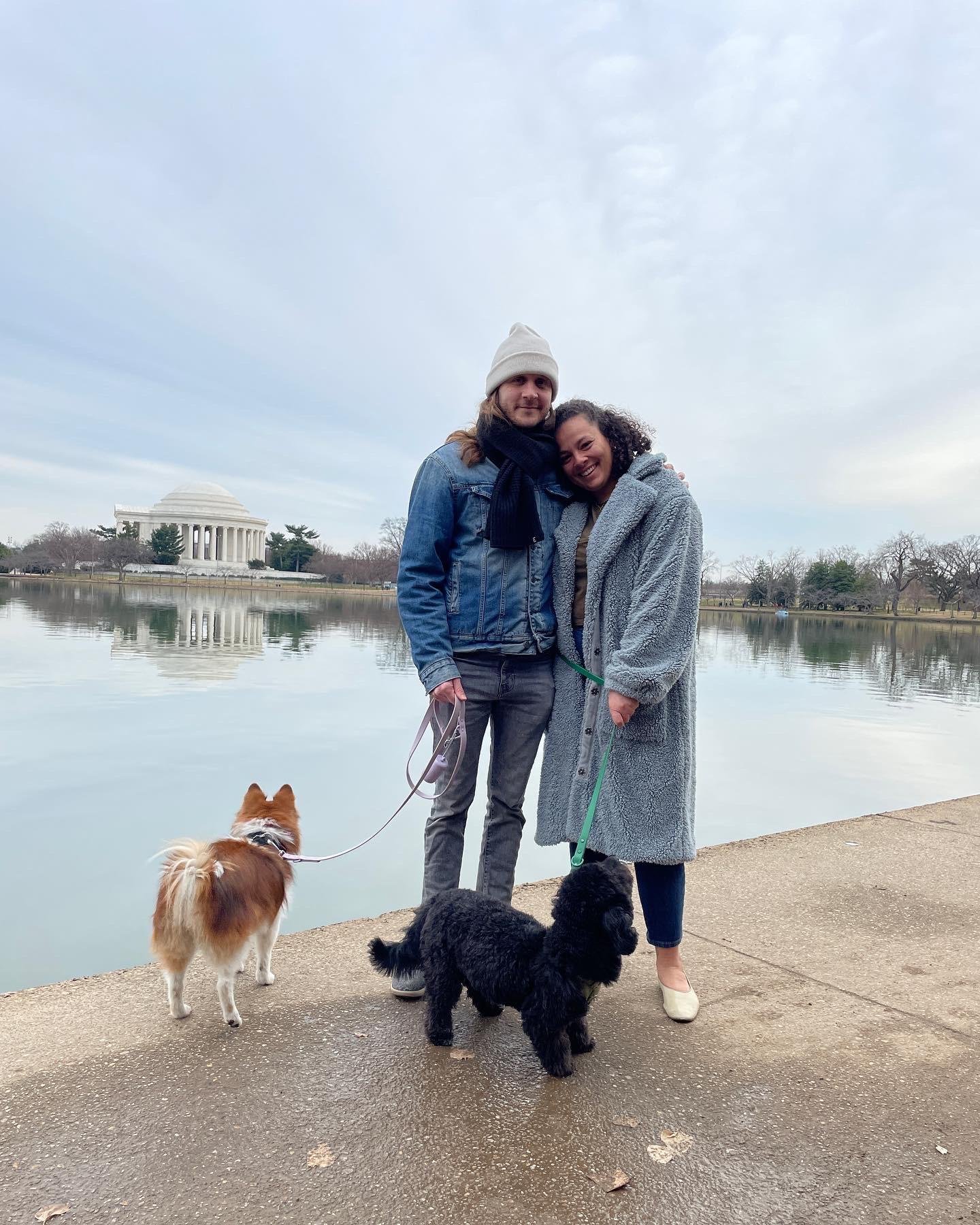 A couple stands with their two dogs in front of the Tidal Basin in Washington, DC. The Jefferson Memorial is in the background. 