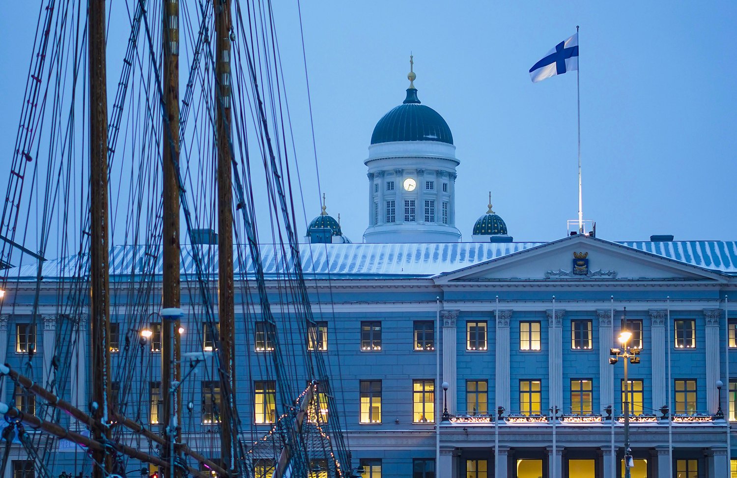 En kvällsbild av Helsingfors stadshus där det lyser i fönstren. På taket vajar Finlands flagga och bakom stadshuset syns domkyrkans torn. I förgrunden ser man masterna från en segelbåt.