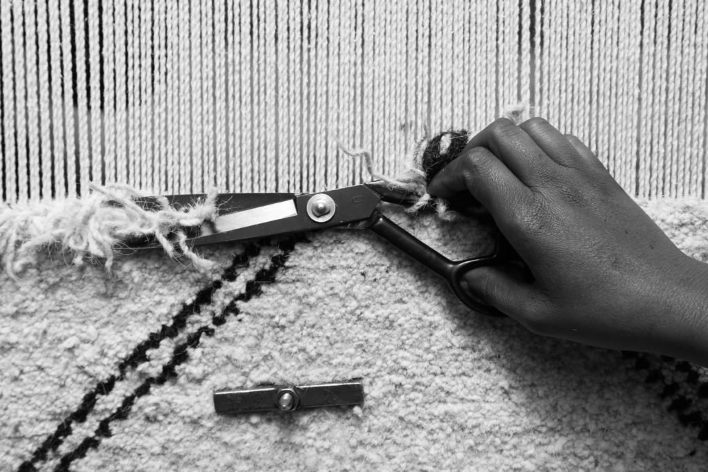 A black and white photograph of a rug weaver trimming the wool with scissors.
