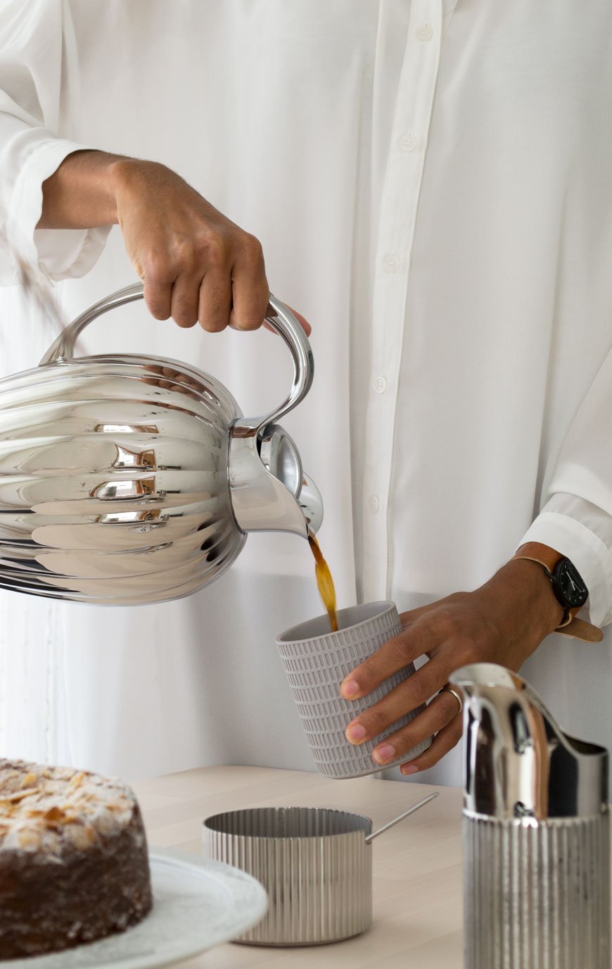 A woman pouring coffee from a stainless steel thermo into a grey ceramic beaker.