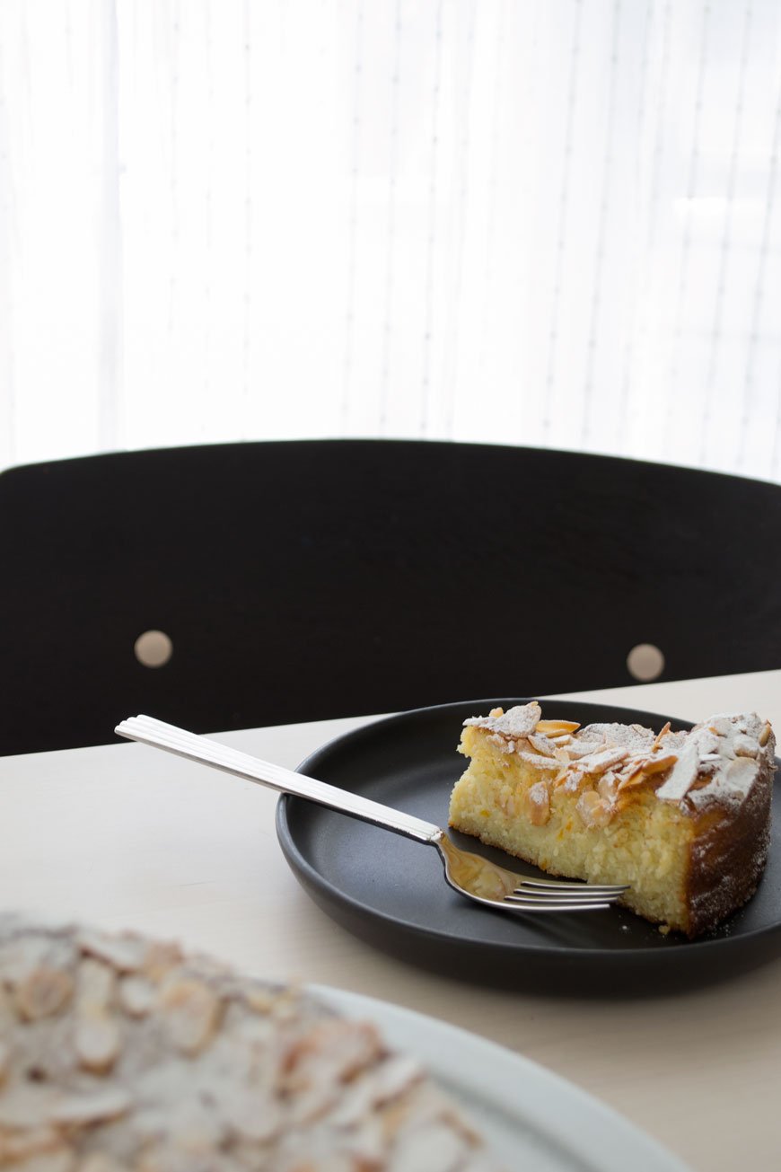 A slice of orange and almond cake styled on a black plate with a stainless steel Bernadotte fork.