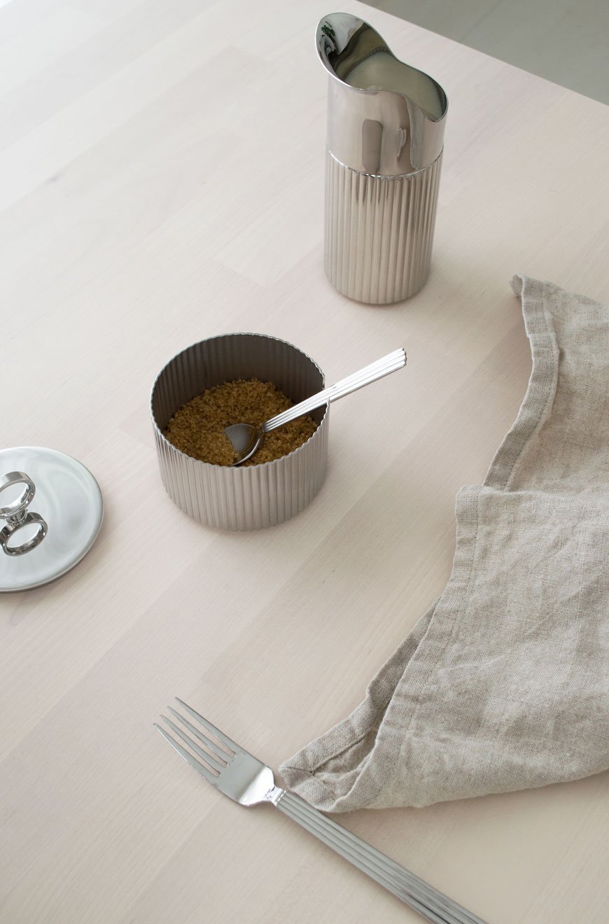 Minimalist styling of a ridged stainless steel milk jug and sugar pot with natural linen napkin. 