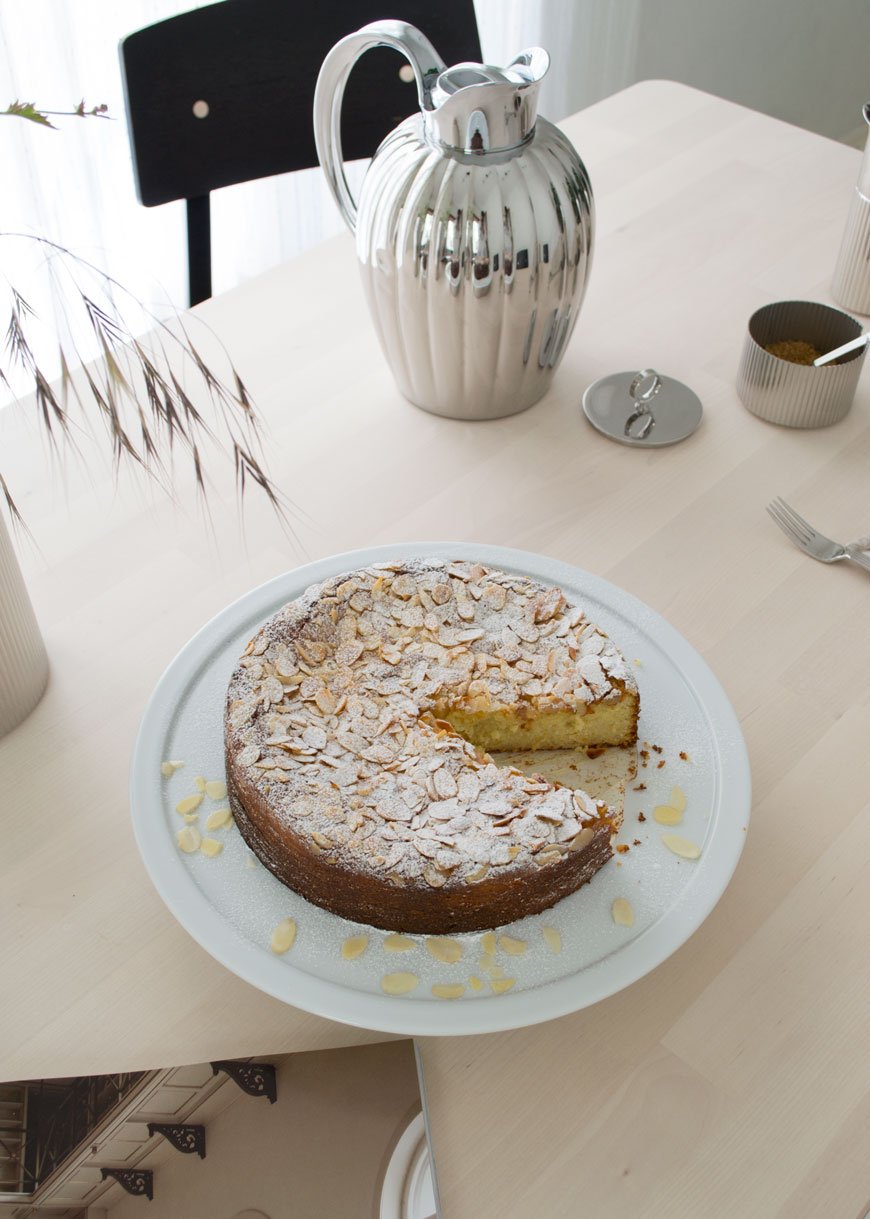 Looking down onto an orange and almond cake on a white serving plate with a thermo of coffee in the background