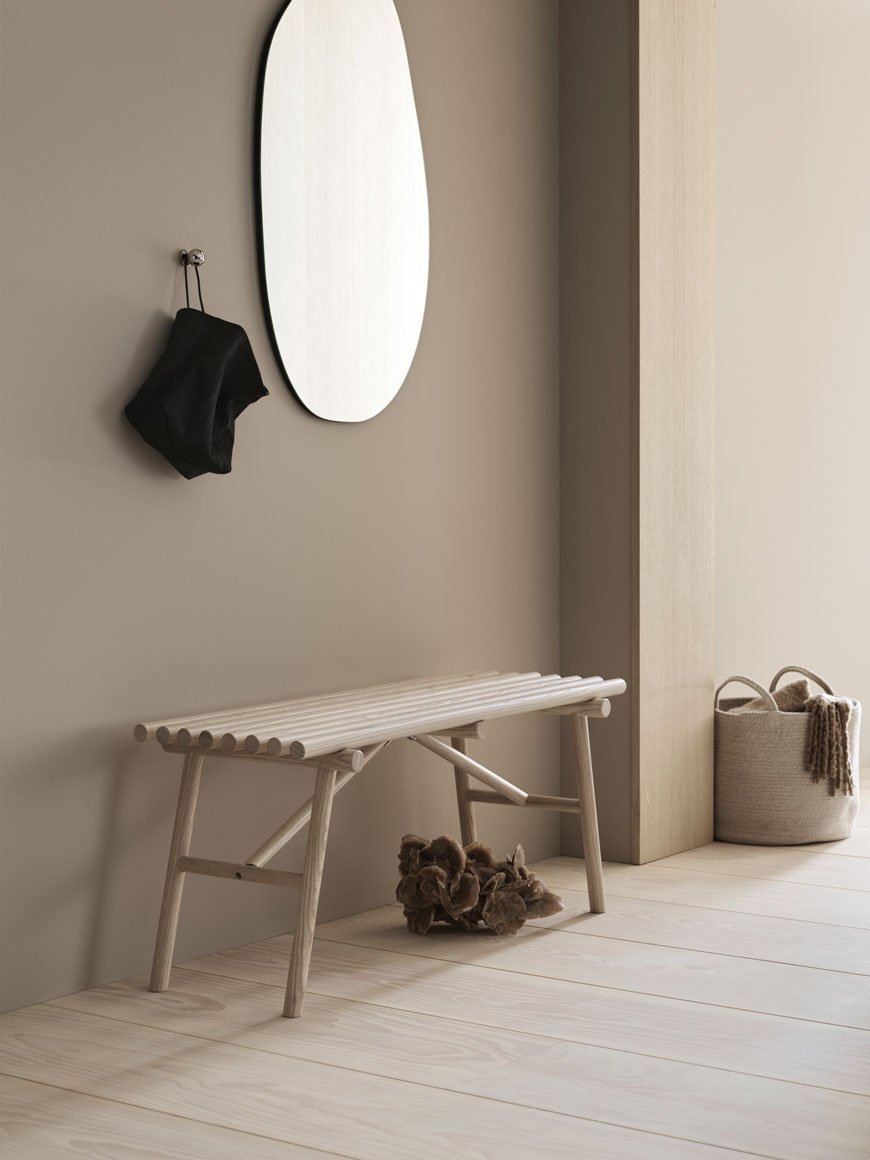 A pale ash wood bench sits underneath an organically shaped mirror for H&M Home. 