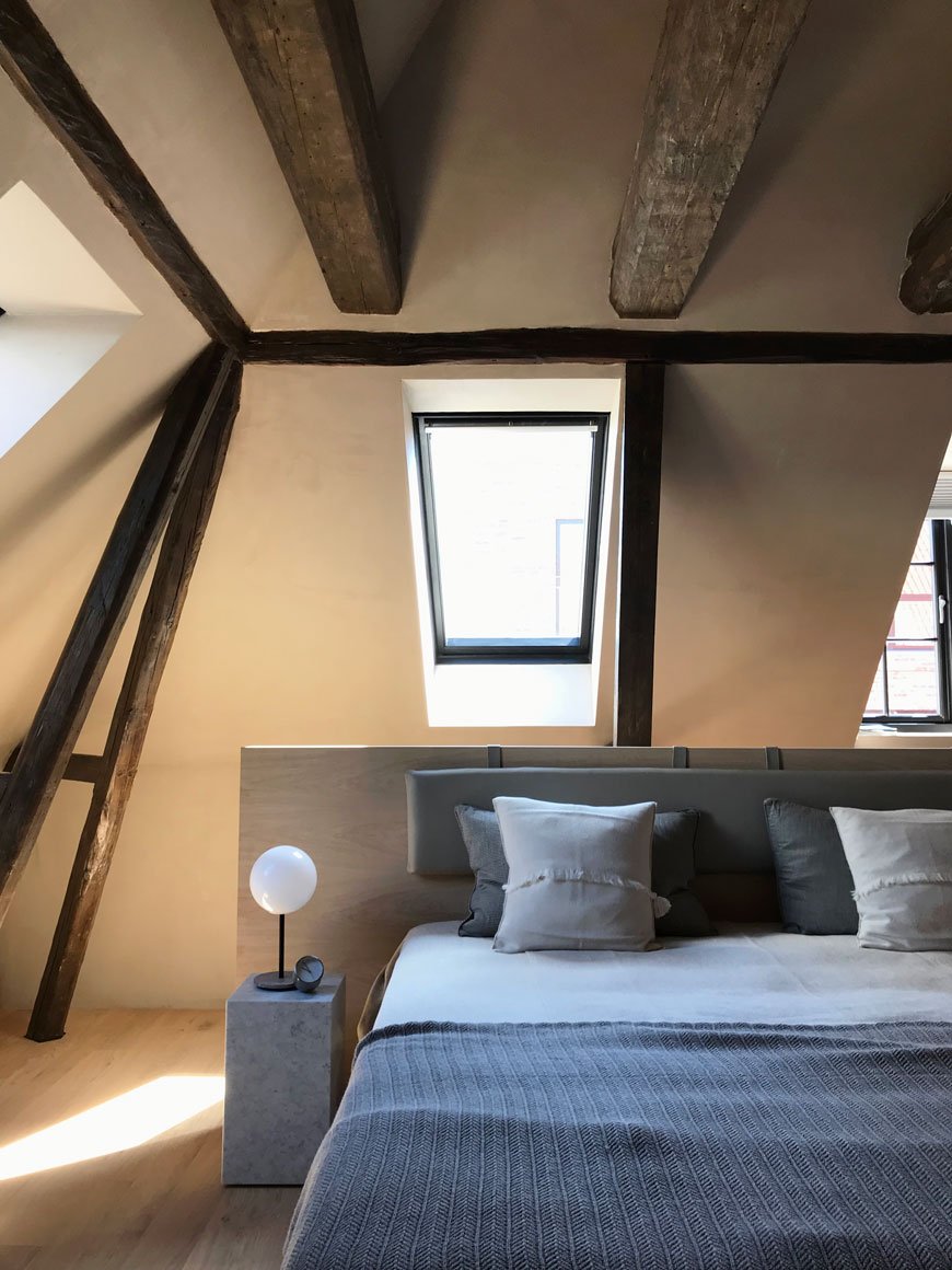 A light filled, cosy loft bedroom suite  with exposed timber beams dressed in blue and grey bedlinen at The Audo residence in Copenhagen. 