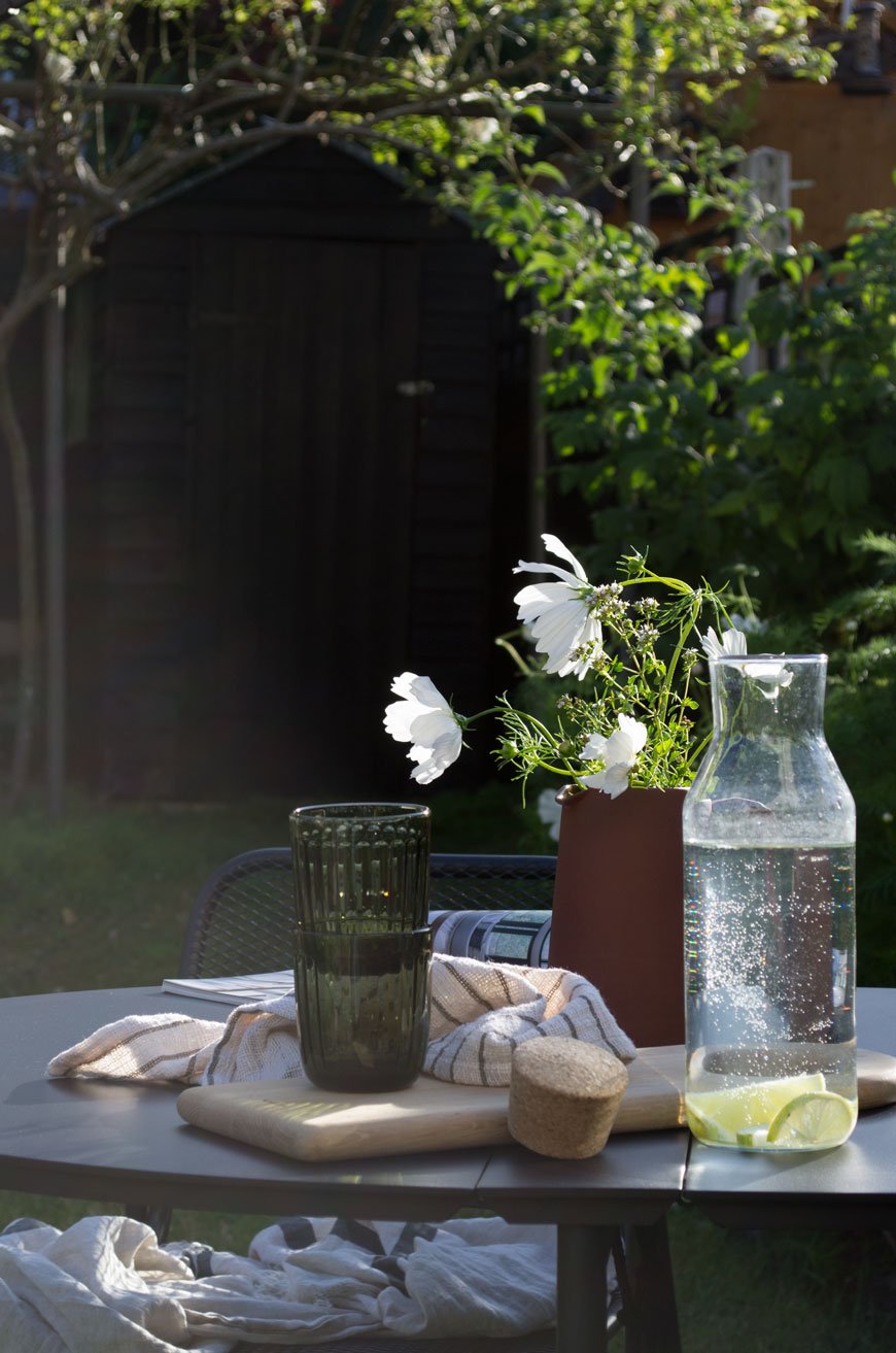 A light drenched table in the garden set with glasses, a pitcher full of white flowers and linen napkins. 