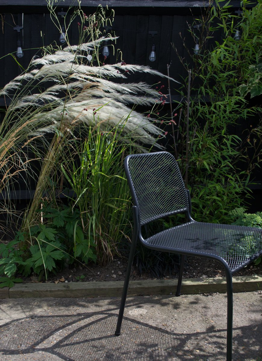 Black Mira outdoor chair with mesh seat by Skagerak sitting in front of an ornamental grass border. 