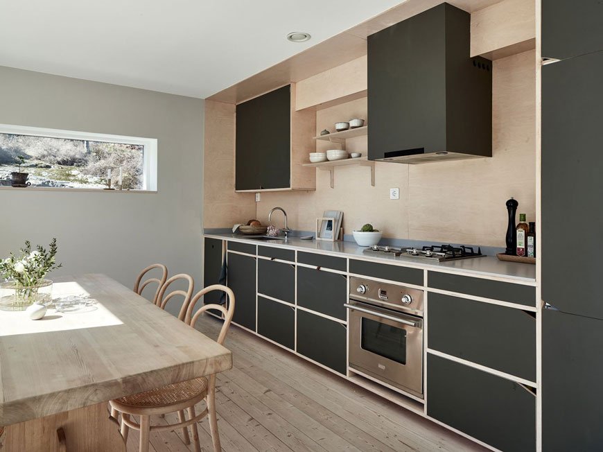 A Scandinavian open plan kitchen dining space with a plywood splashback and dark green kitchen cabinets.
